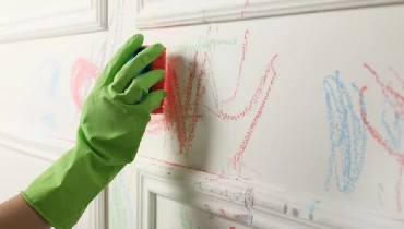 Person wearing rubber gloves and using a sponge to remove crayon from a white wall.