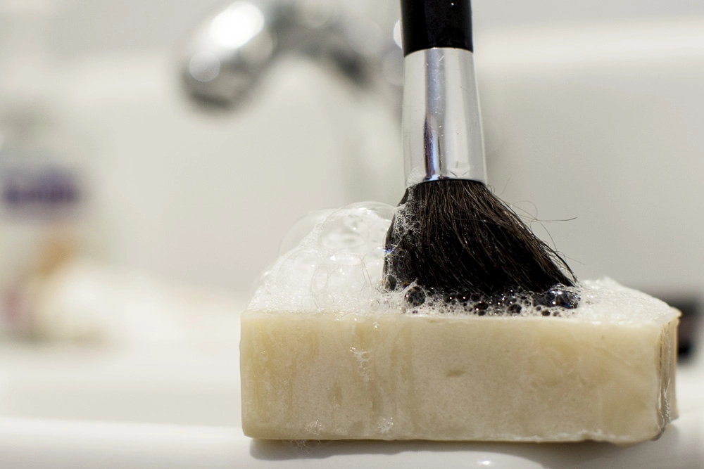 The bristles of a makeup brush on a bar of soap, with a blurry sink faucet in the background