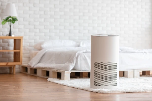 air purifier in foreground with cozy white bedroom in background
