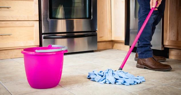 A person's hands using a mop to clean a kitchen floor, with a pink bucket nearby