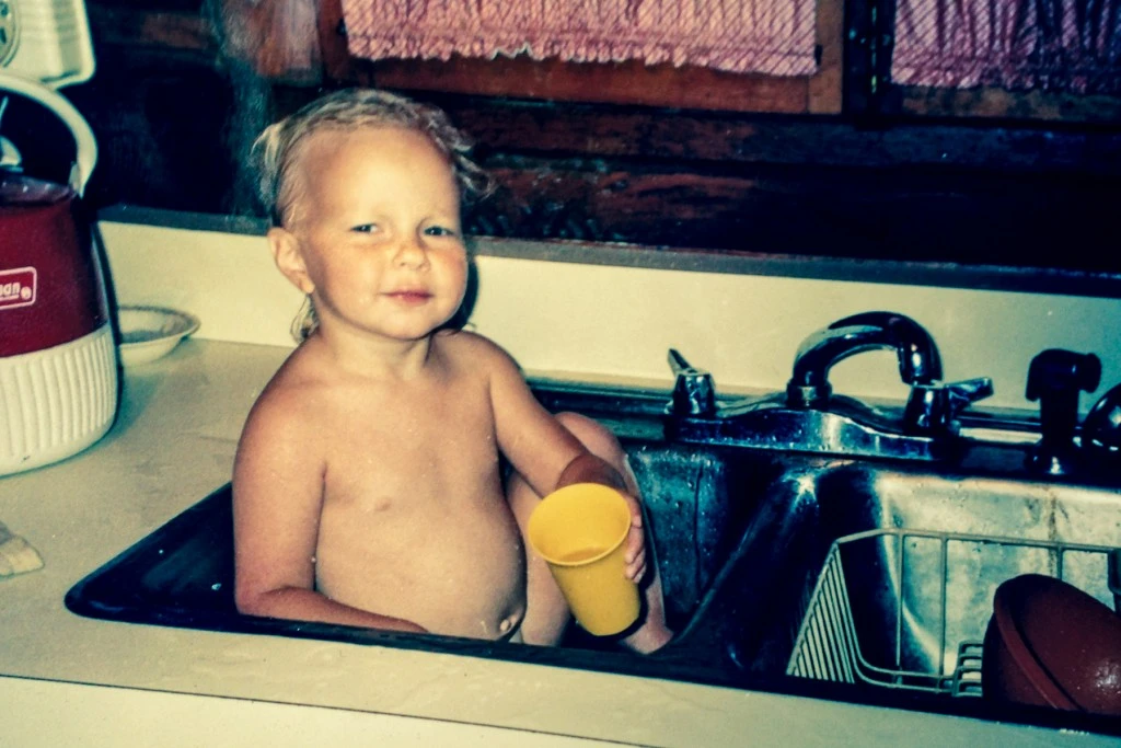 A young child sitting in a kitchen sink and holding a yellow cup