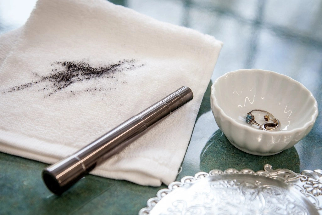 Black mascara stain on a white towel next to a tube of mascara and jewelry dish