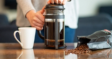 Person holding a French press full of coffee on a table, next to a white mug and bag of spilled coffee beans.