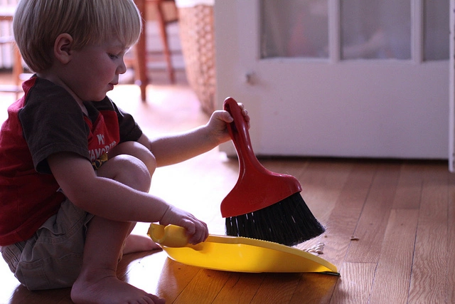 A toddler crouched on the floor using a small brush and dustpan to sweep. Photo cred: Jessica Lucia on Flickr.