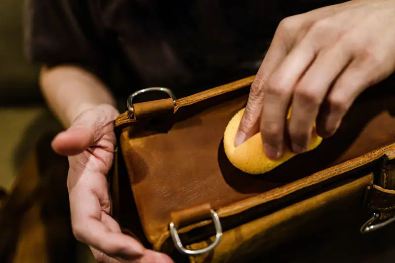 Hand with yellow soft sponge cleaning stains on the surface of a brown leather bag.