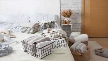 Mesh metal containers full of clothes and linens on the bed and floor in a bedroom. Cup of coffee on a serving tray on the bed.