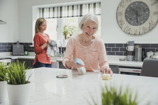 Elderly women cleaning kitchen counter with help from her daughter in the background