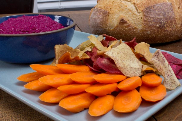 A gray serving tray with beet hummus, carrots, and veggie chips in front of a loaf of bread
