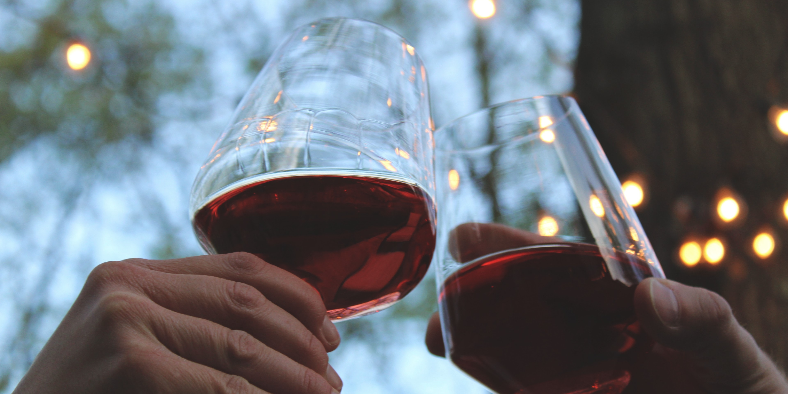 Two wine glasses containing red wine being clinked together in an outdoor setting, photo by rikkia hughes on Unsplash