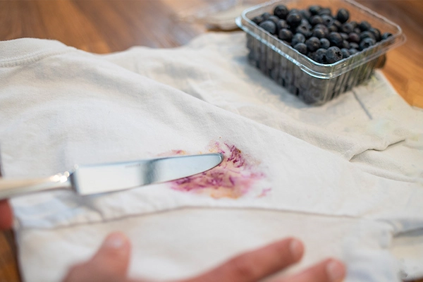 Berry stain on white cloth napkin with silver knife and pint of blueberries spread out on table.