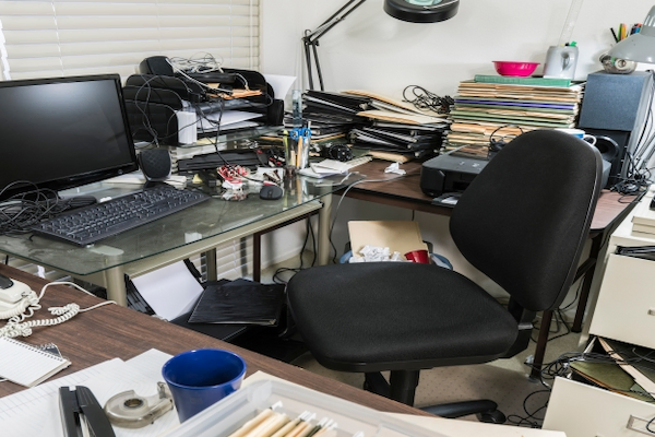 Desk of a home office covered in files and surrounded by clutter.