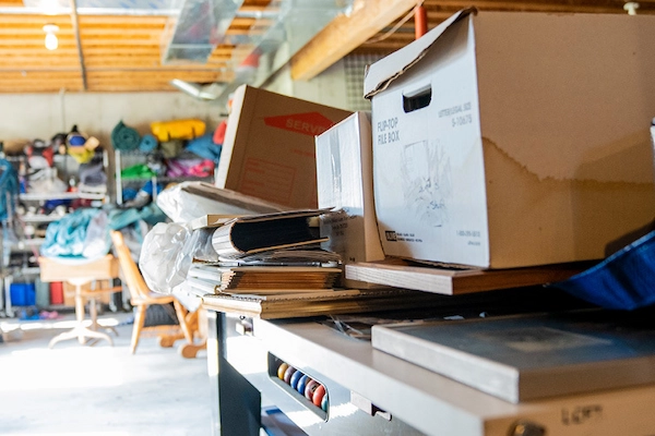 Carboard boxes and miscellaneous items piled on table and shelves inside cluttered garage.