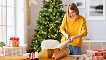 Happy young woman wrapping holiday gifts with Christmas tree in background.
