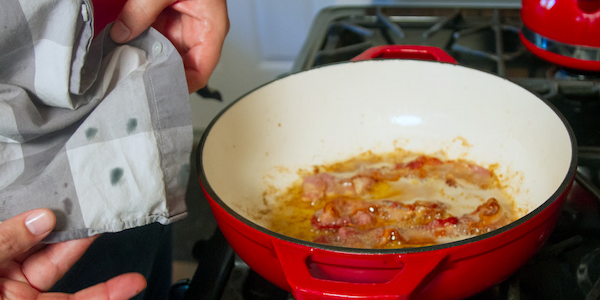 Close-up of gray checkered shirt with grease stains held up beside red pan with bacon grease on stovetop.