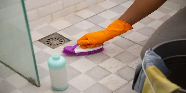 Person wearing orange rubber glove scrubbing shower floor with brush.