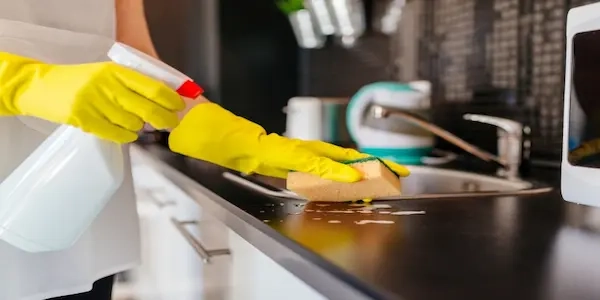 Person wearing yellow rubber gloves cleaning black countertop with spray and yellow sponge.