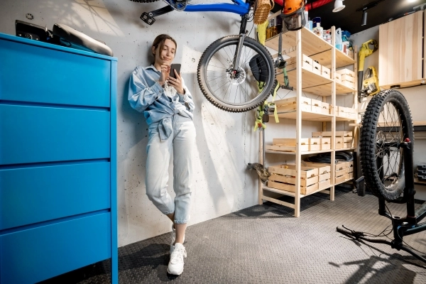 Young woman on her phone standing in a garage with bicycle hanging from ceiling.