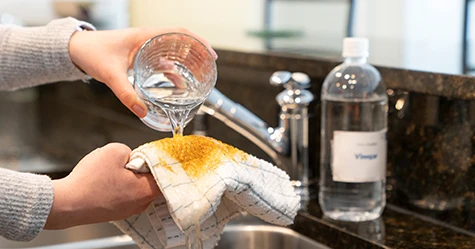 Water being poured from a glass onto a white cloth stained with turmeric, with a bottle of vinegar in the background